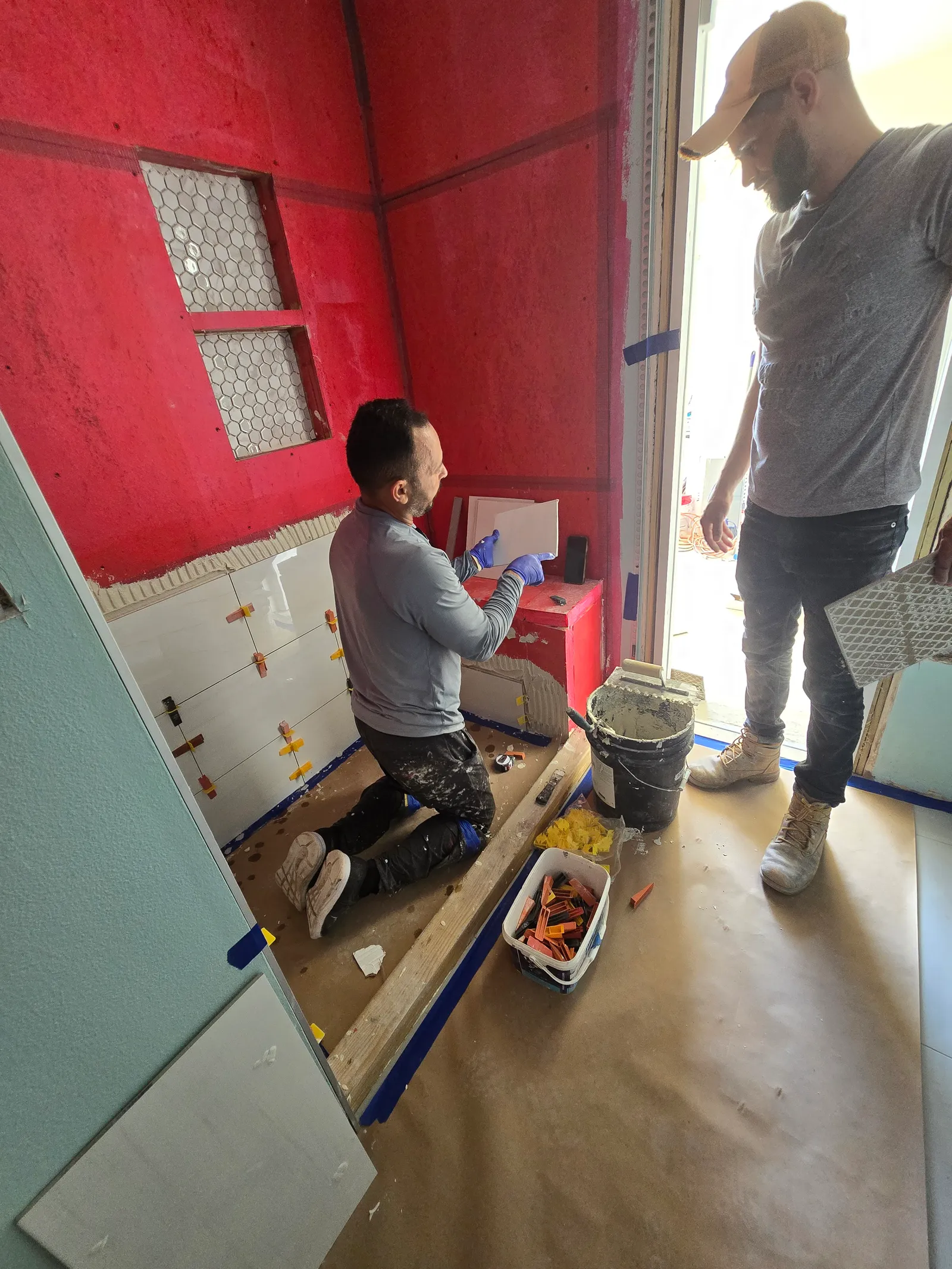 Two crew members setting wall tile over red waterproofing membrane in a bathroom under renovation