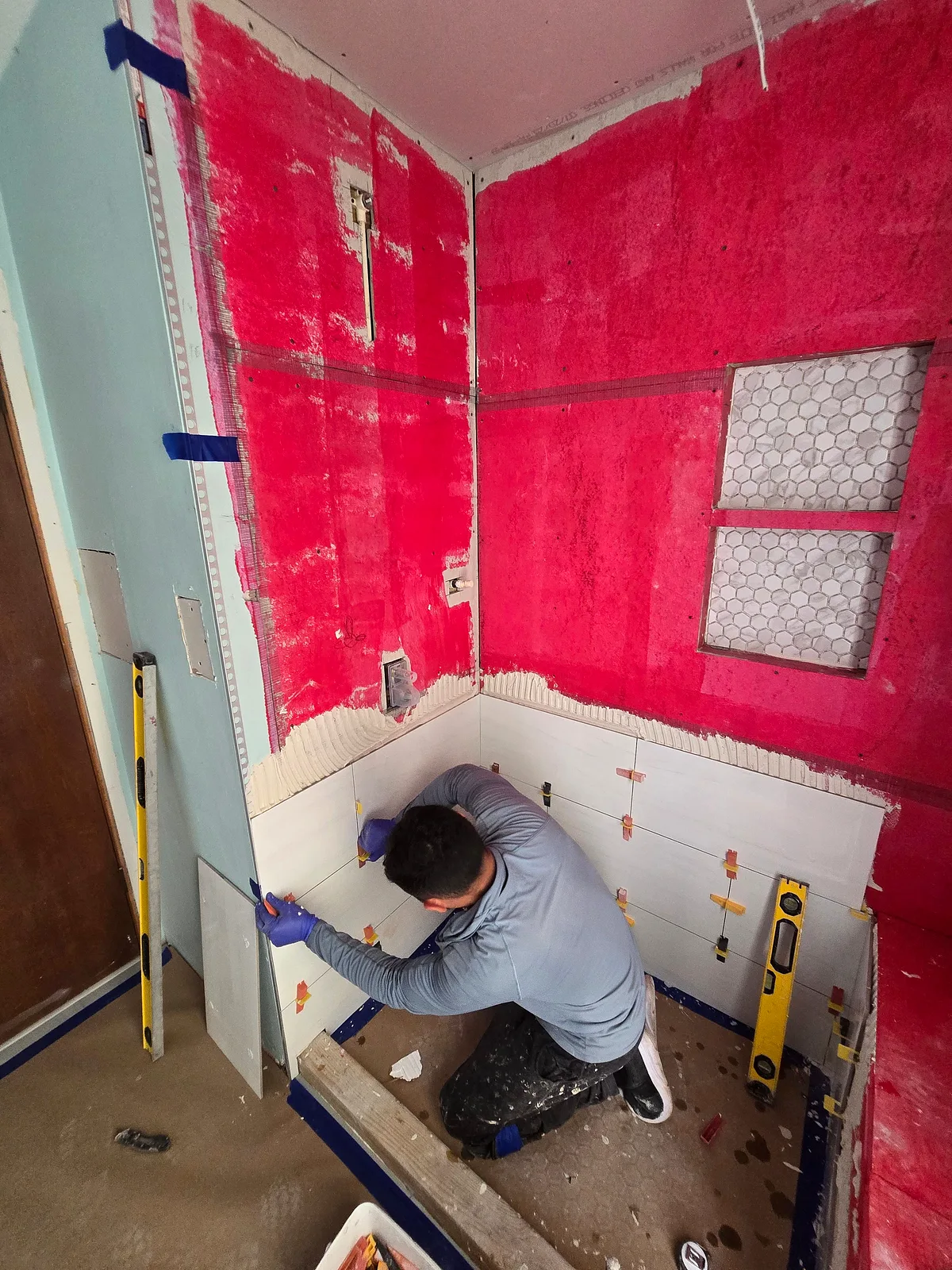 Crew member kneeling to set wall tile over red waterproofing membrane in a bathroom under renovation